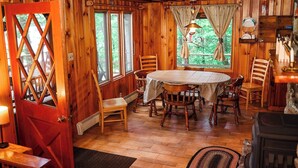 A wooden cabin interior with a dining table surrounded by chairs, a fireplace, and large windows offering a view of the outdoors. - Gramps Camp • Moose Pond (Bridgton)