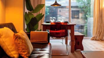 Un salon chaleureux avec un canapé en cuir, une table à manger avec des chaises, une grande plante et une vue sur l’extérieur.