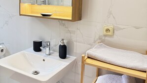A bathroom with a marble wall, a wooden medicine cabinet, a white sink, a black soap dispenser, and a towel rack with folded towels. - Riverside Apartment 11 (Hlavní město Praha)