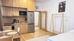 A compact kitchen with wooden cabinets, a microwave, and a refrigerator. There is a dining table with chairs and a bed with a blue bedspread. - Riverside Apartment 11 (Hlavní město Praha)