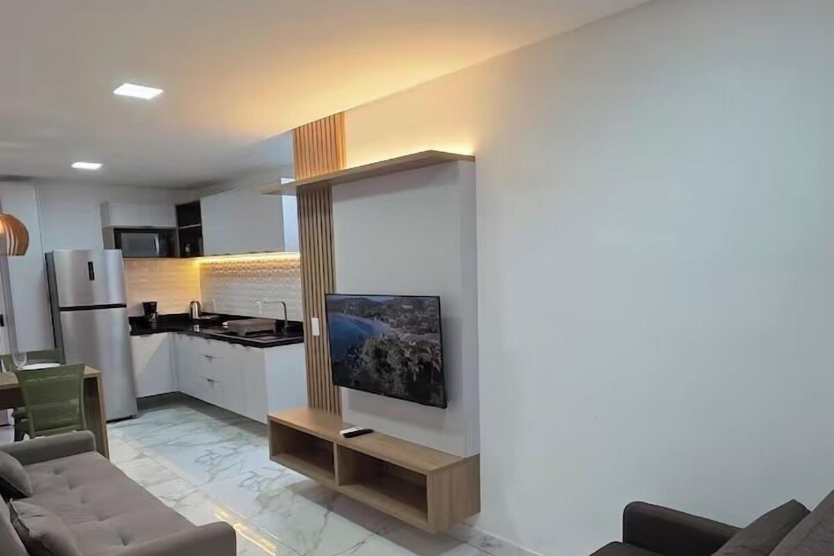 Una sala de estar moderna con un televisor de pantalla plana grande montado en la pared, un sofá y un comedor con una cocina al fondo.