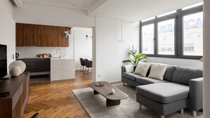 A modern living room with a grey sectional sofa, a wooden herringbone floor, a kitchen with wooden cabinets, and a large window with a view of buildings.