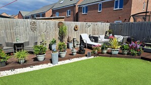Un jardin avec une pelouse, des plantes en pot, une terrasse en bois et du mobilier d’extérieur.