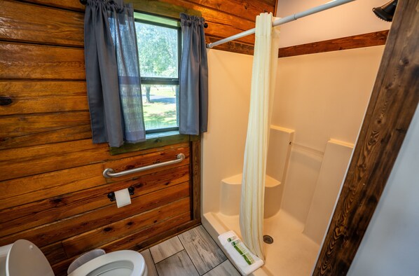 A wooden cabin bathroom with a toilet, a shower, and a window with curtains. - Fishtale Lodge (Buchanan)
