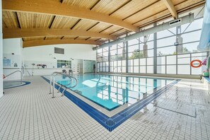 Una piscina interior con techo de madera, ventanas amplias y suelo de baldosas.