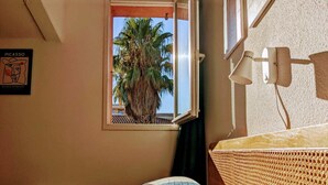 Una habitación con cama, una ventana que da a una palmera, una lámpara de pared y un cartel del Museo Picasso.