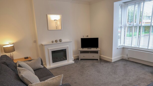A living room with a grey sofa, a fireplace, a television on a stand, and a window with blinds. - Jubilee Two Bedroom Bungalow Pet Free (Penrith)
