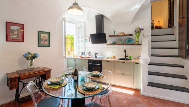 A dining area with a glass table, chairs, and a bottle of wine. There is a kitchen in the background with white cabinets and a black range hood. A staircase is visible to the right. - GuestHost - Charming 100 sqm apartment, accommodating up to 6 people, located on the ground floor of a typical Ligurian building.The property is located in the center of Cervo, one of Italy's most charming villages, in a convenient and well-served locati (Cervo)