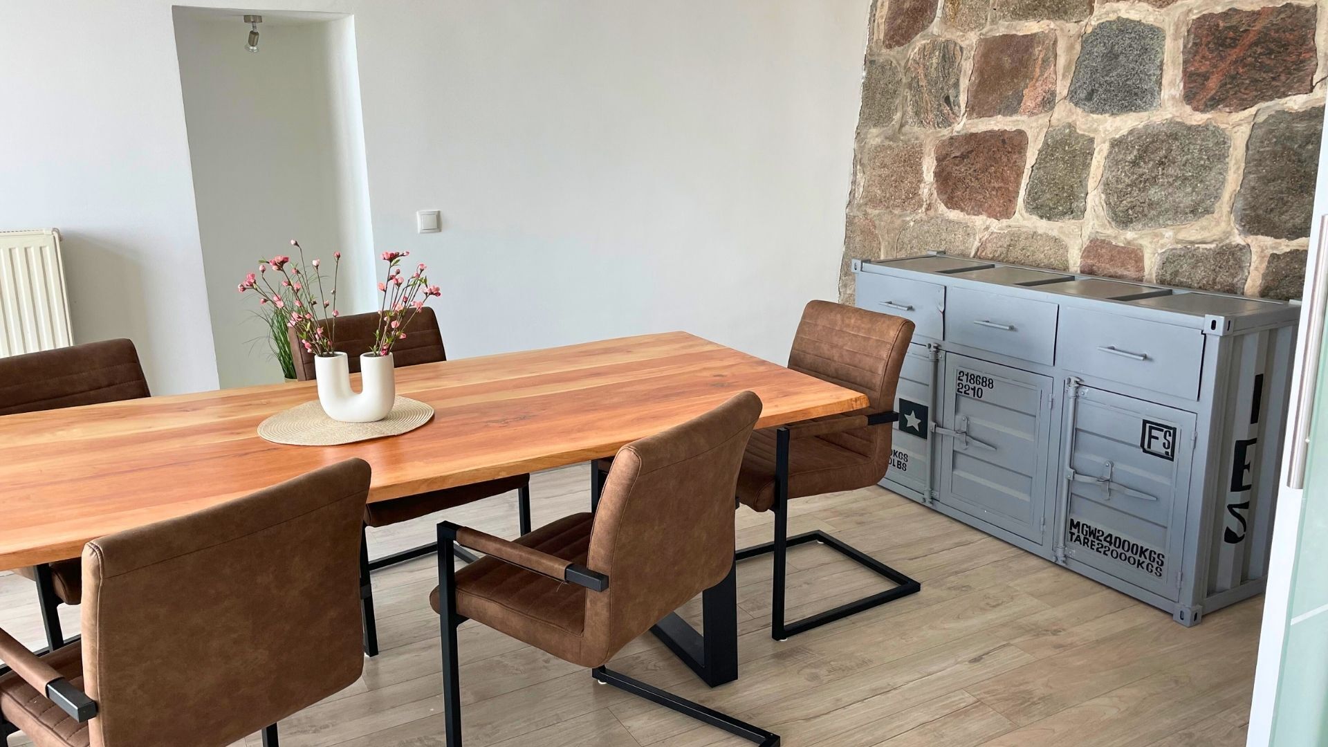 Un coin repas avec une table en bois, des chaises marron, un vase de fleurs et un mur en pierre.