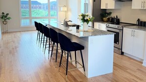 A modern kitchen with a central island, bar stools, and a view of a building through large windows. - Luxurious House with pond view and Jacuzzi (Oakville)