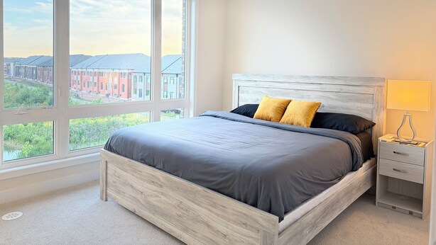 A bedroom with a large window, a bed with grey bedding, a wooden headboard, and a bedside table with a lamp. - Luxurious House with pond view and Jacuzzi (Oakville)