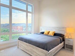 A bedroom with a large window, a bed with grey bedding, a wooden headboard, and a bedside table with a lamp. - Luxurious House with pond view and Jacuzzi (Oakville)