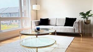 Uma sala de estar moderna com um sofá, duas mesas de vidro, um abajur e vista para um edifício através de grandes janelas.