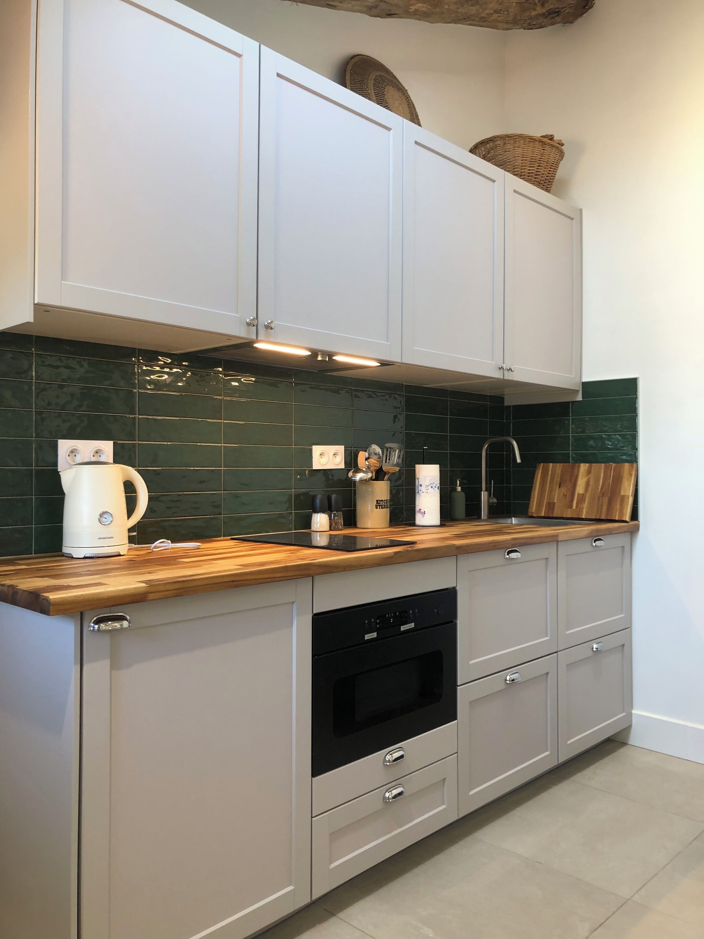 Une cuisine moderne avec des meubles de couleur gris clair, un plan de travail en bois, un four encastré et un crédence recouverte de carreaux verts.