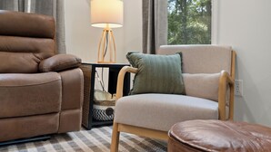 Ein Wohnzimmer mit braunem Ledersofa, einem beigen Sessel, einem Holztisch mit Lampe und Blick auf Bäume durch ein Fenster.