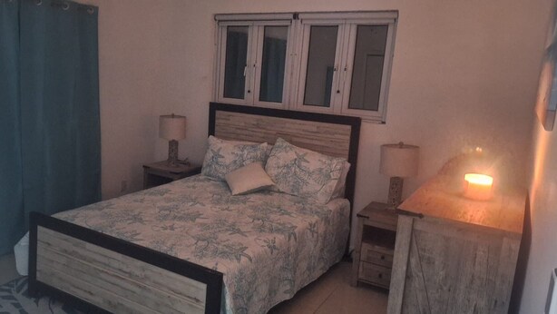 A bedroom with a bed, a nightstand, a lamp, a window, and a curtain. - Charming cottage in Exuma for a relaxing getaway. Come and enjoy Hideaway Cove (Exuma)
