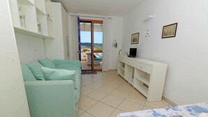 A room with a light green sofa, a bed with a patterned bedspread, a white console table with a TV, and a sliding glass door leading to an outdoor area with a view. - Relax in Leuca - Terrace with Panoramic Sea View - Casa Michele (Santa Maria di Leuca)