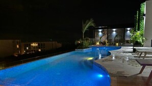 A nighttime swimming pool with a curved edge, surrounded by a tiled deck and some greenery.