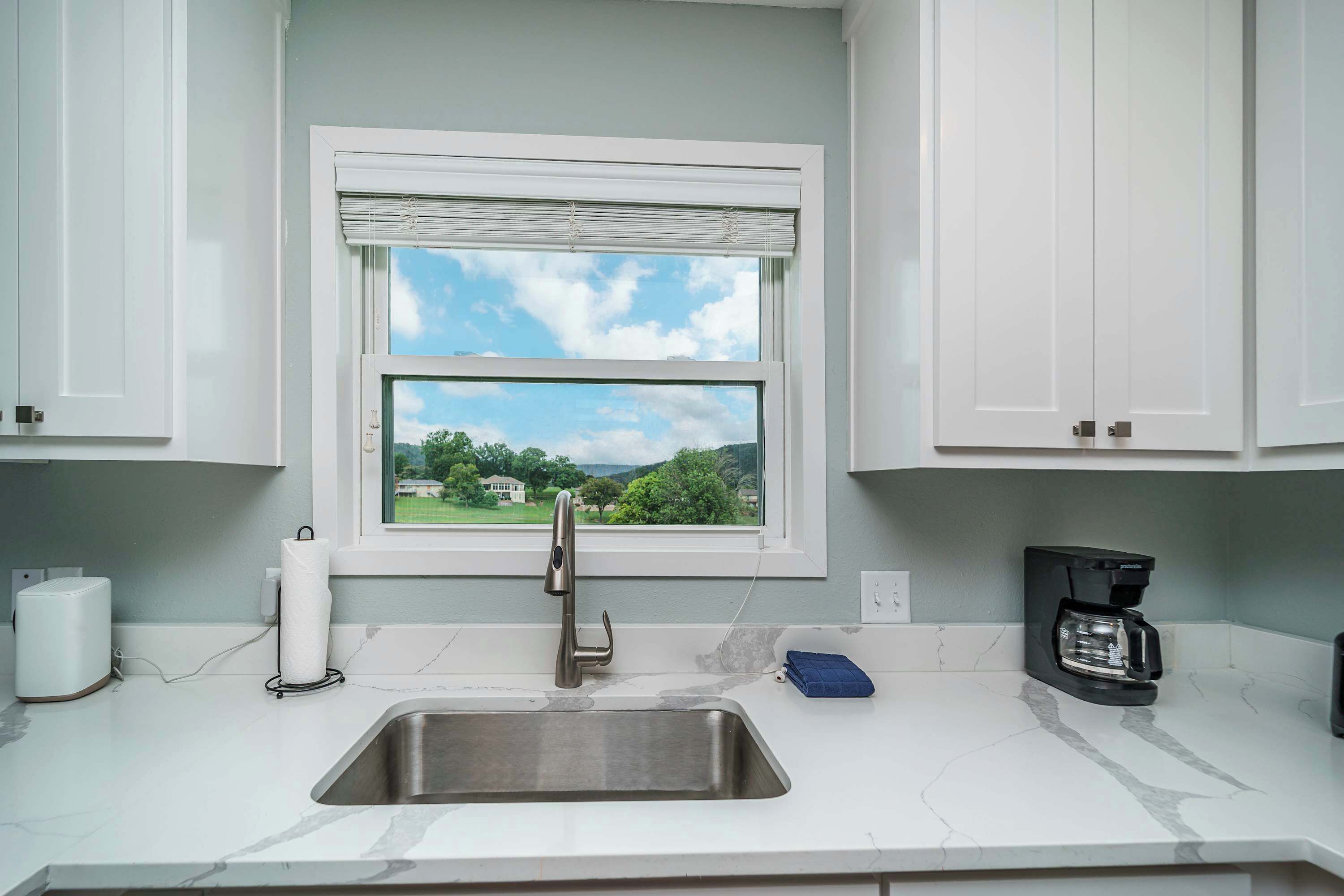 A kitchen with a stainless steel sink, a window with a view of greenery, a coffee maker, and a paper towel holder.