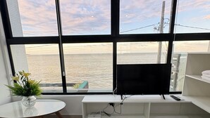 A modern living room with a large window overlooking a body of water, a flat-screen TV mounted on a white console, and a grey sofa. - Melhores Flats - Deslumbrante  (Joao Pessoa)