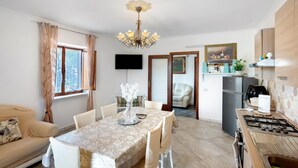 Een eethoek met een tafel voor vier personen, een bank, een aan de muur bevestigde televisie, en een keuken met een fornuis en een koelkast.