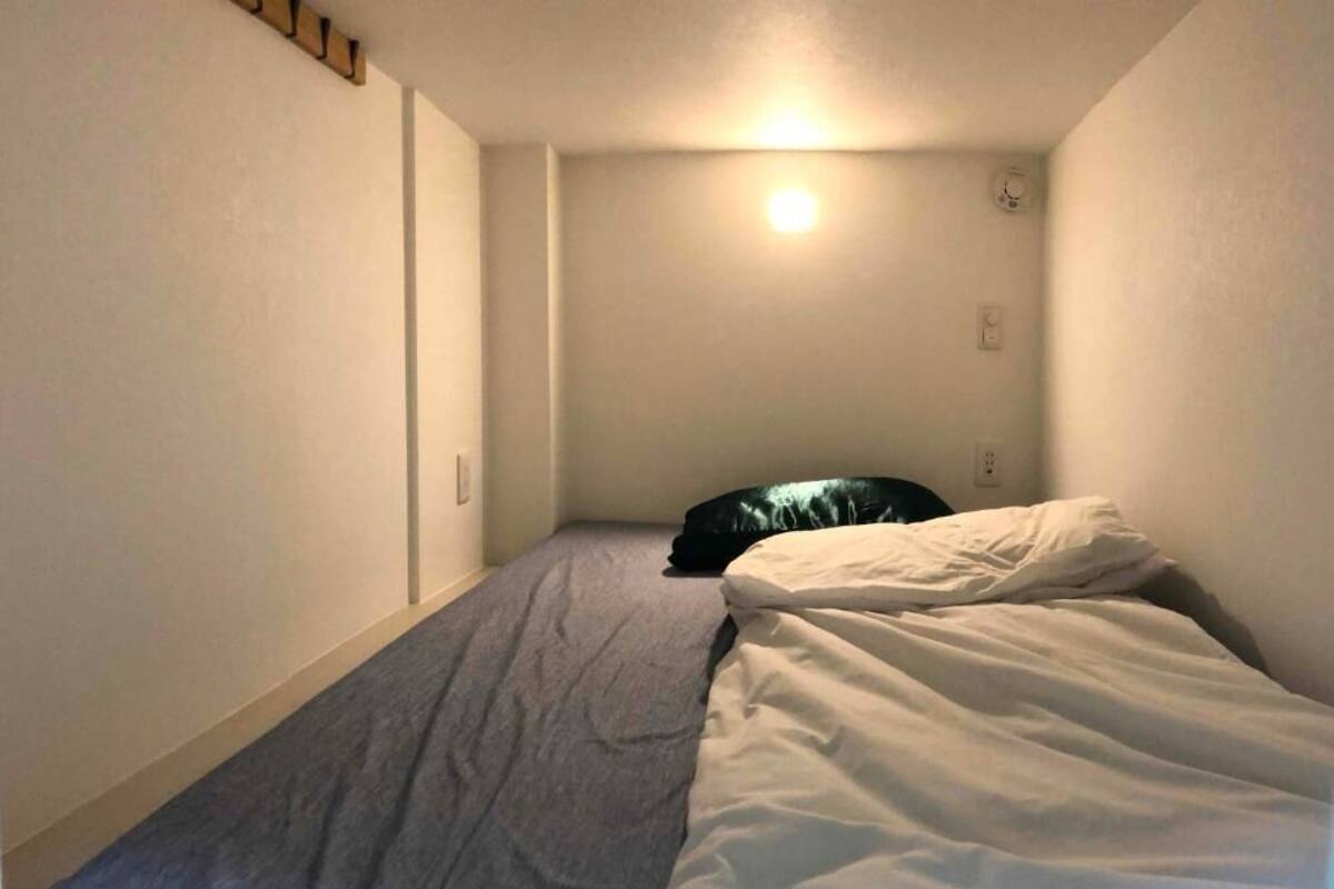 Quarto pequeno com uma cama de solteiro, parede simples e luminária no teto.