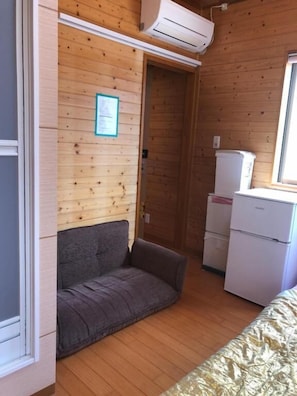 A compact room with wooden walls, a grey sofa, a small kitchenette with a fridge, and a window. - Guest house Wa N Wa 2 (Osaka)