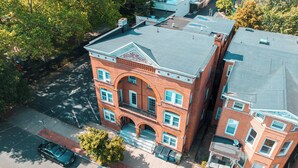 A multi-story brick building with a central arched entrance, adjacent smaller buildings, and a parking area with two cars. - A brick's throw away to Sally's Apizza! (New Haven)