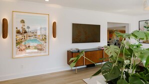 Una sala de estar moderna con un cuadro grande enmarco de una escena tropical, una consola de madera y un cuadro abstracto de gran tamaño.