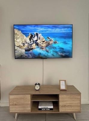 A flat-screen TV mounted on a wall above a wooden TV stand with a clock and books.