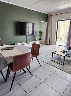A modern living room with a dining area, a flat-screen TV mounted on the wall, a large window with curtains, and a small side table.