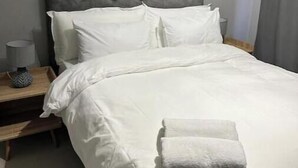 Una cama bien hecha con sábanas blancas y una toalla doblada, una lámpara de noche y una mesita de madera.