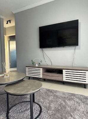 Ein modernes Wohnzimmer mit einem Wand-Fernseher, einer Holzunterhaltungsecke darunter und einem runden Couchtisch auf einem grauen Teppich.