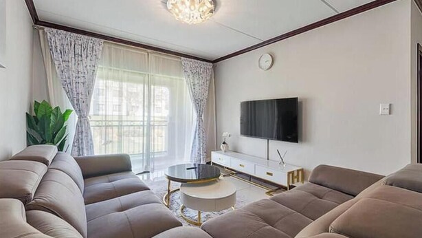 A modern living room with a sectional sofa, a round coffee table, and a flat-screen TV mounted on the wall. - Gem in Fourways Modern 2-Bed Apartment (Randburg)