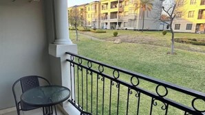 A balcony with two chairs and a table, overlooking a well-maintained lawn and a row of yellow buildings. - Gem in Fourways Modern 2-Bed Apartment (Randburg)