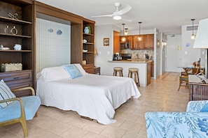 A bedroom with a bed, a wooden wardrobe, a small kitchen area, and a dining table with chairs. - Oceanfront Maui Escape! Panoramic Views, Pool, Hot Tub & Steps to Beach (Lahaina)