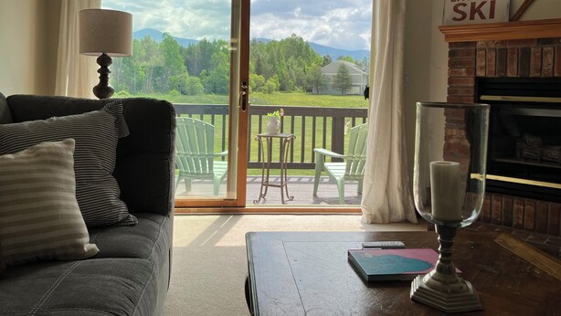 A living room with a sofa, a table with a book and a candle, a fireplace, and a view of a green landscape through a glass door.