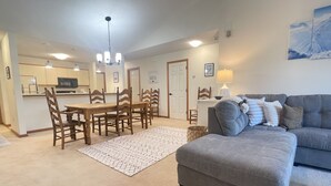 A living room with a grey sofa, wooden dining table, and chairs. There is a kitchen area with cabinets and a microwave.