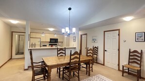 A spacious dining area with a wooden table and chairs, a kitchen in the background, and a ceiling light fixture.