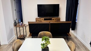 A conference room with a white table, four chairs, a potted plant, a television on a wooden stand, and a small table with a bottle and a remote control.