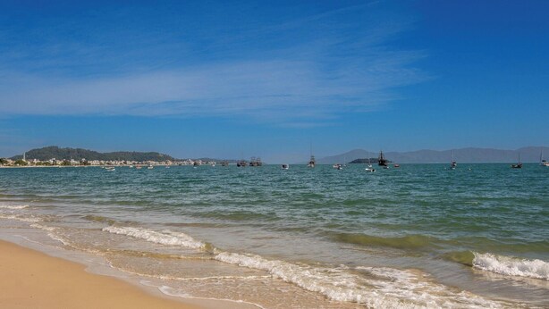 A beach with calm waters, boats, and distant hills. - Encantador Apto Beira-mar Floripa Rpb037 (Florianópolis)