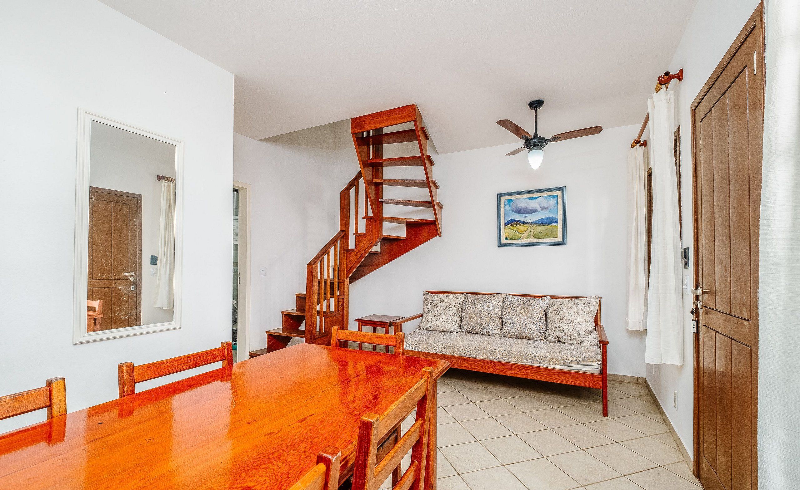 A dining area with a wooden table and chairs, a wooden staircase, a ceiling fan, and a sofa with patterned cushions.