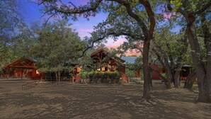 Un edificio de estilo cabaña rústica rodeado de árboles, con suelo de tierra y un banco al frente.