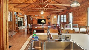 Interior rústico de una cabaña con piso de madera, chimenea, comedor y cocina con pileta y encimera.