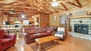 Amplia sala de estar con techo de madera, muebles de cuero, chimenea de piedra y cocina al fondo.