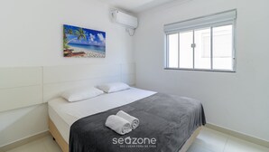 A bedroom with a bed, white headboard, a painting of a beach, an air conditioning unit, and a window with blinds.