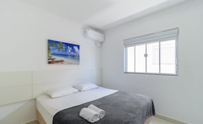Een slaapkamer met een bed, een wit hoofdeinde, een schilderij van een strand, een airconditioner en een raam met zonwering.