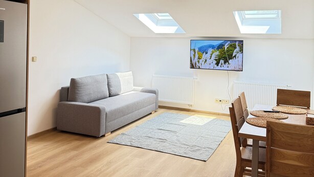 A modern apartment with a grey sofa, wooden dining table, and a built-in kitchen area.
