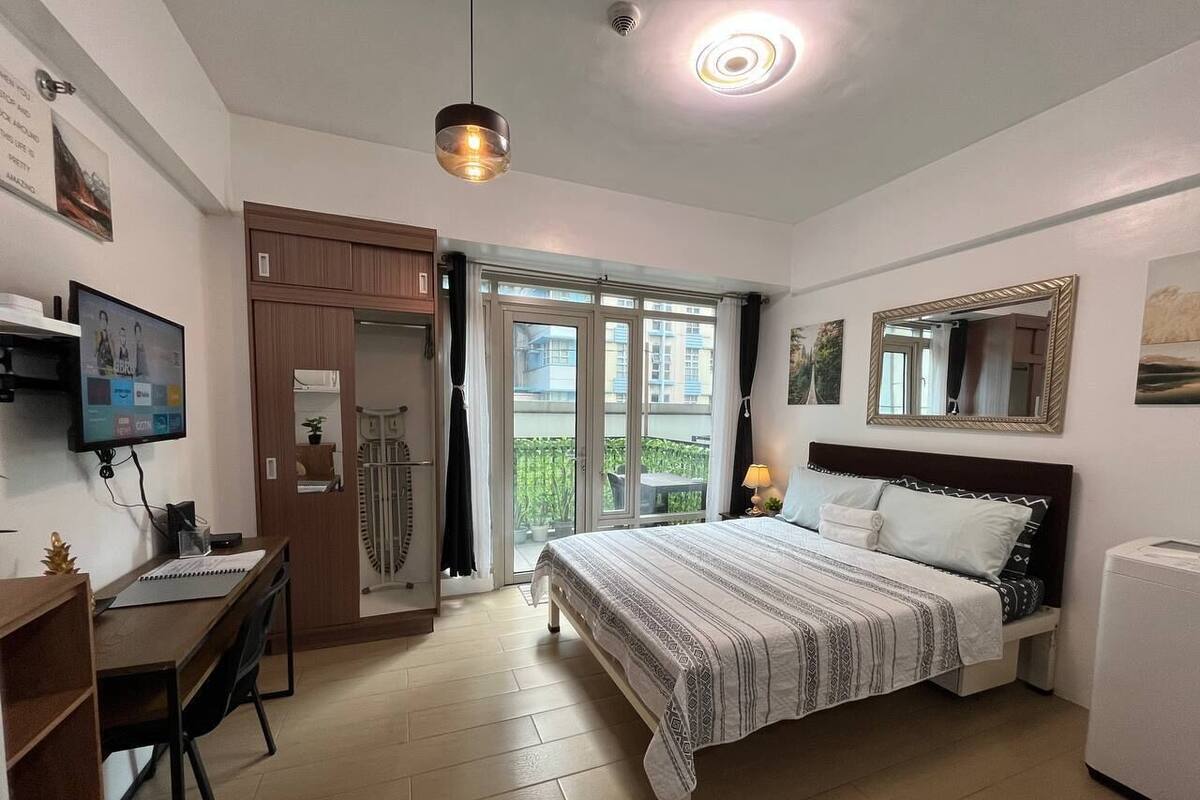 Un dormitorio con cama, escritorio, silla, armario, espejo y ventana con vista a edificios.