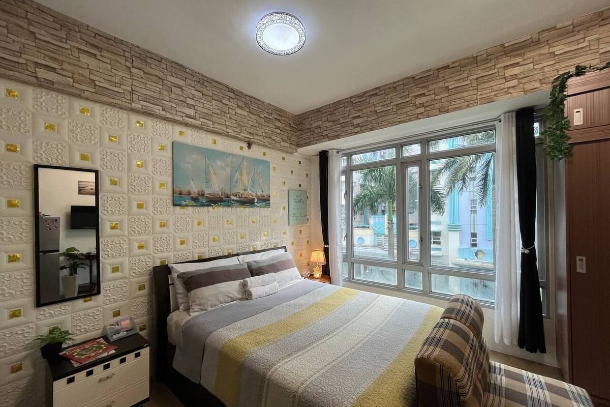 Un dormitorio con cama, mesita de noche, espejo, ventana y vista a palmeras, además de un panel decorativo en la pared.